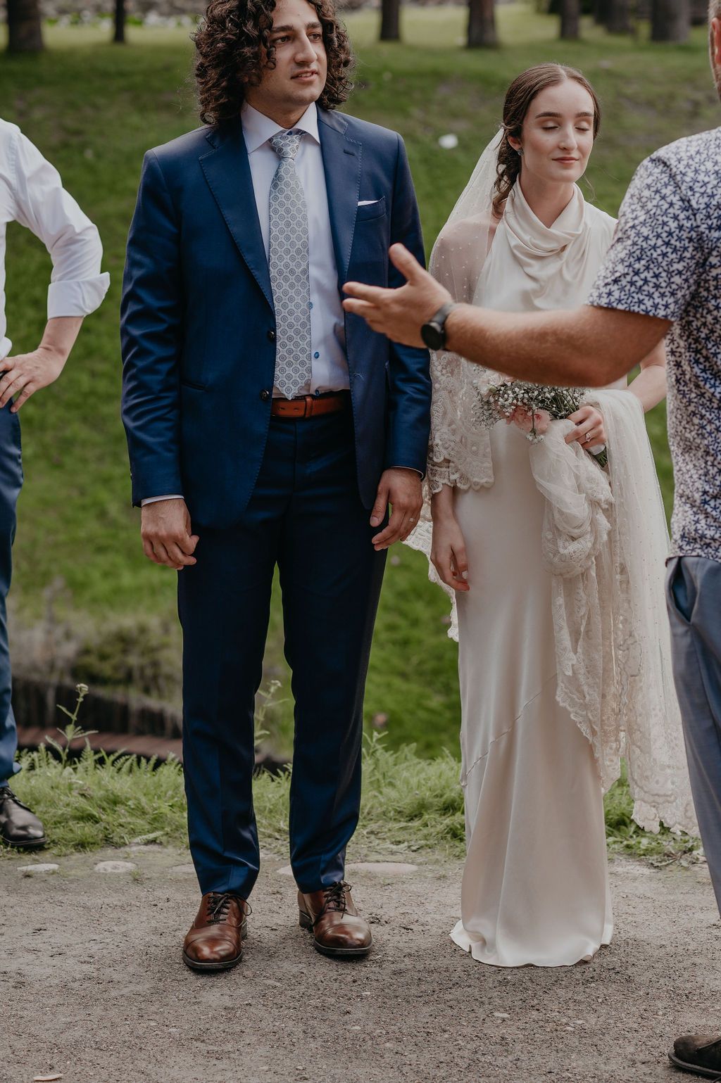 Bride in white gown with veil and groom in blue suit standing outdoors at wedding ceremony.