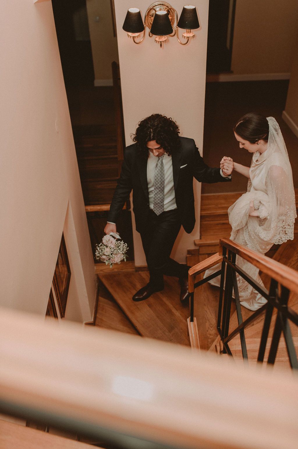 Groom leads bride down elegant wooden staircase, holding hands and bouquet, in classic black suit and white veil.