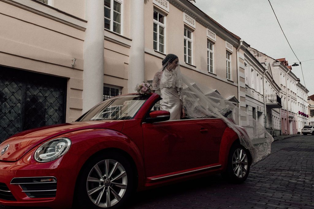 Bride in white gown standing beside red VW Beetle convertible on city street.