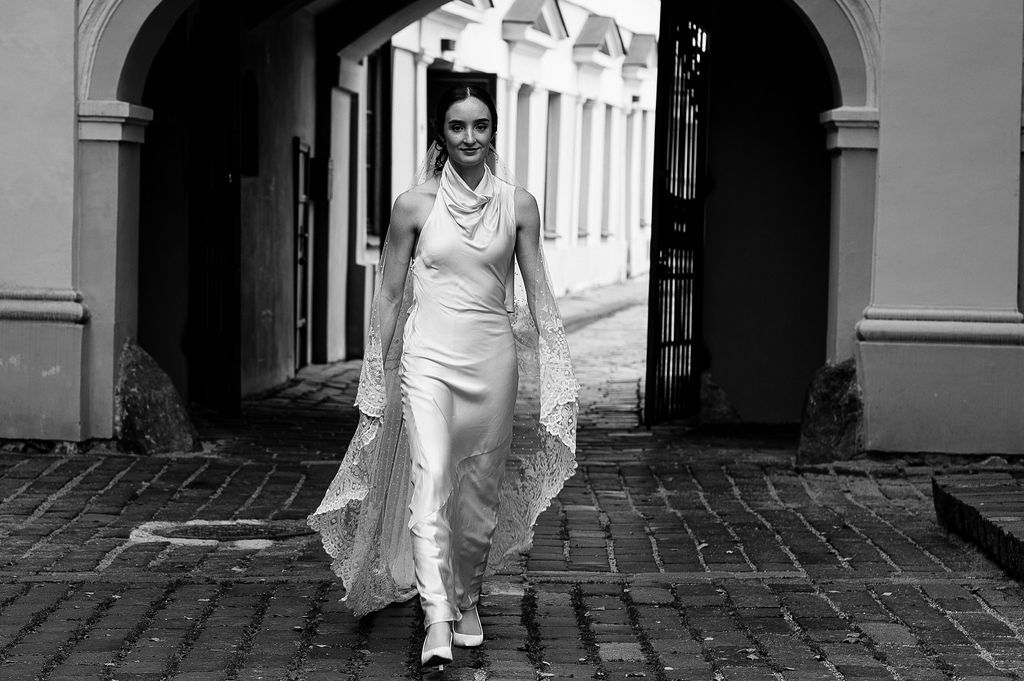 Bride in elegant white satin gown and lace veil walks cobblestone street in black and white photo.