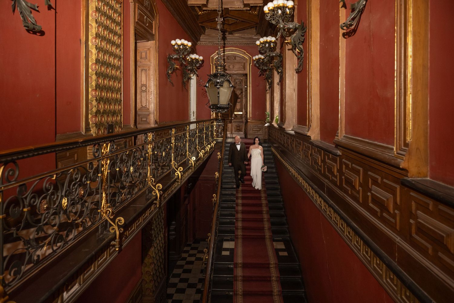 Ornate red hallway with gilded wrought iron railings, chandeliers, and checkered marble floors in a grand historic palace.