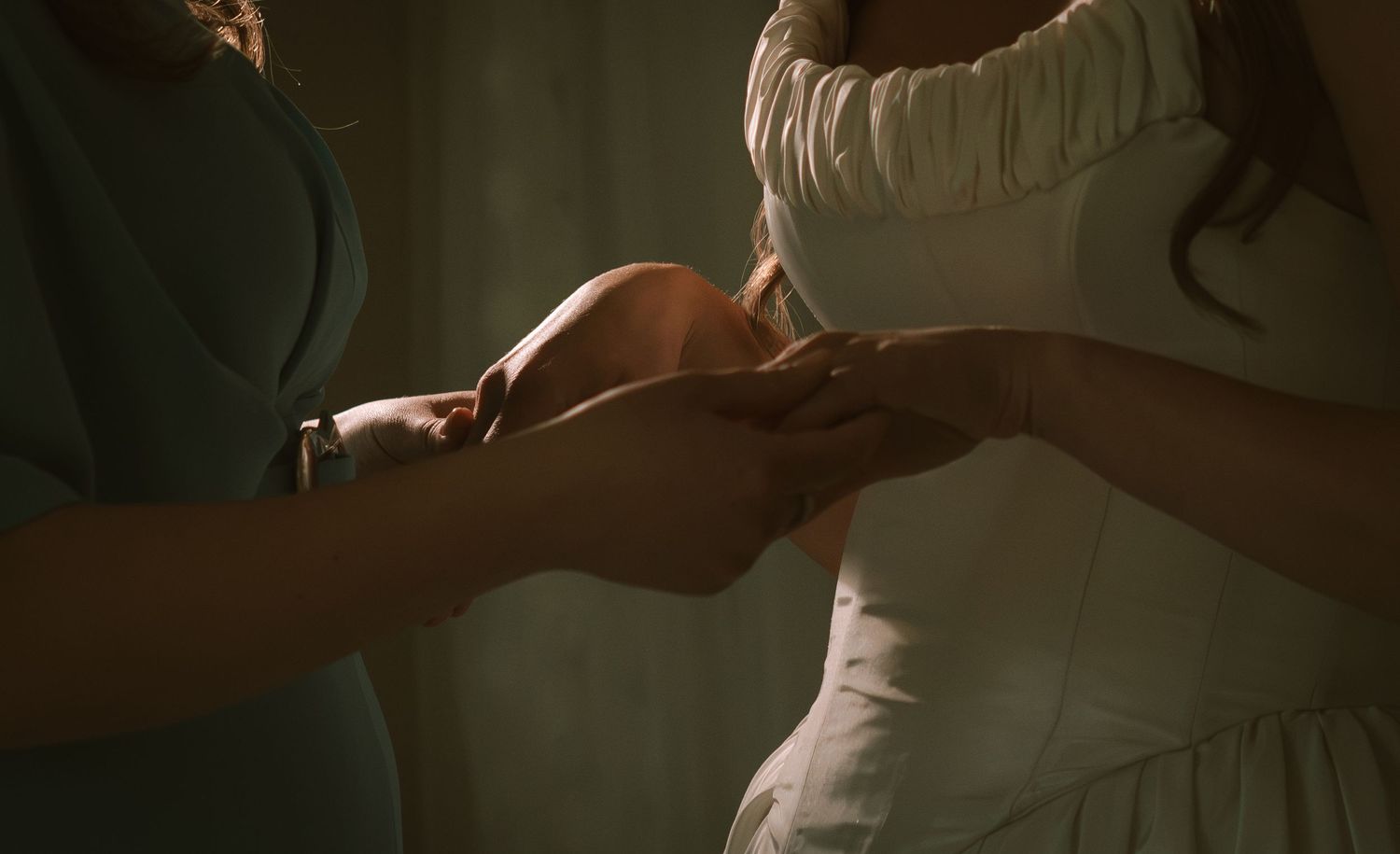 Two women holding hands in an intimate moment, one wearing a white dress, in a softly lit setting.