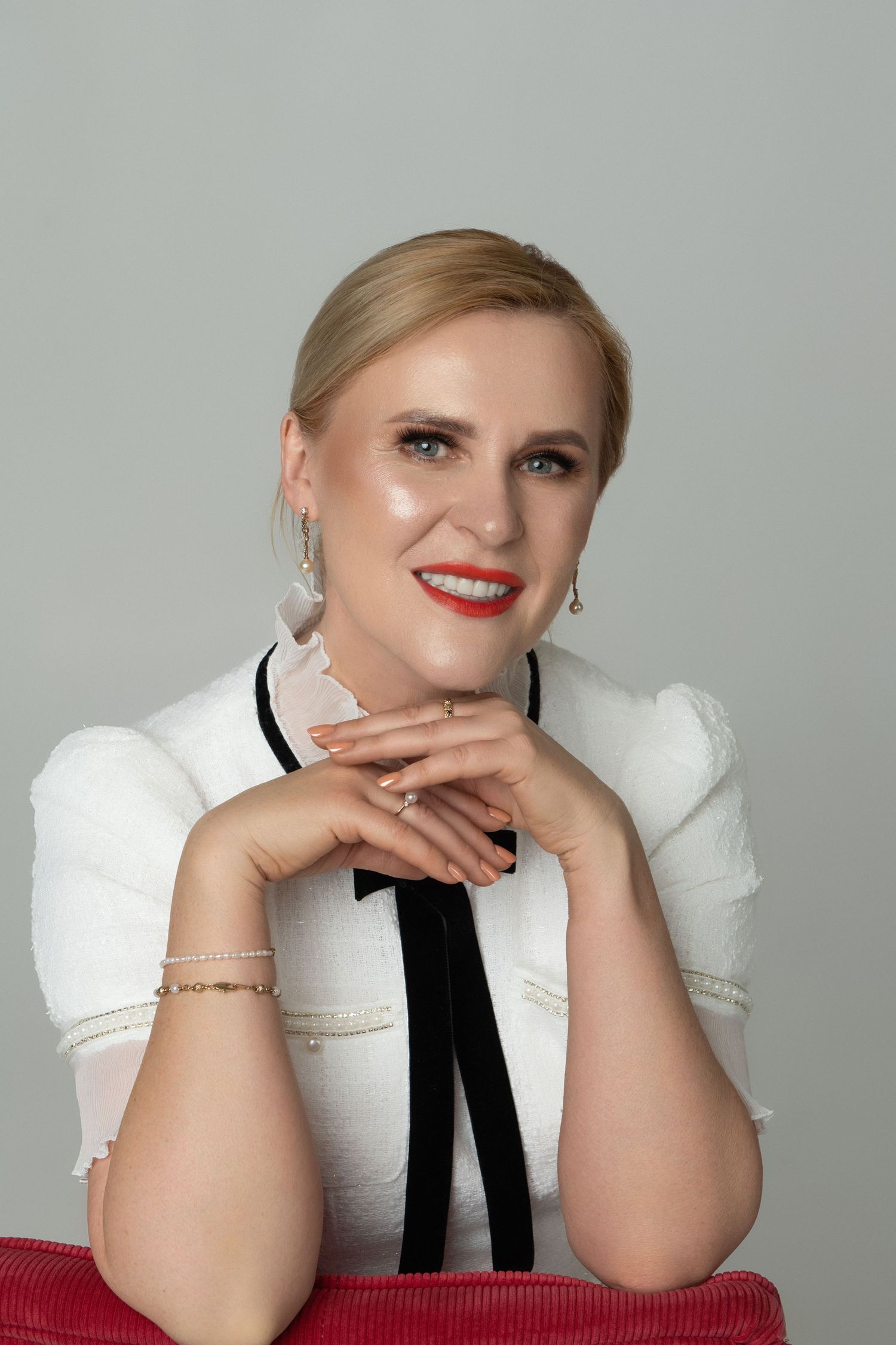 Smiling blonde woman in white blouse with black bow tie, red lips, drop earrings, bracelet, posing against gray background.