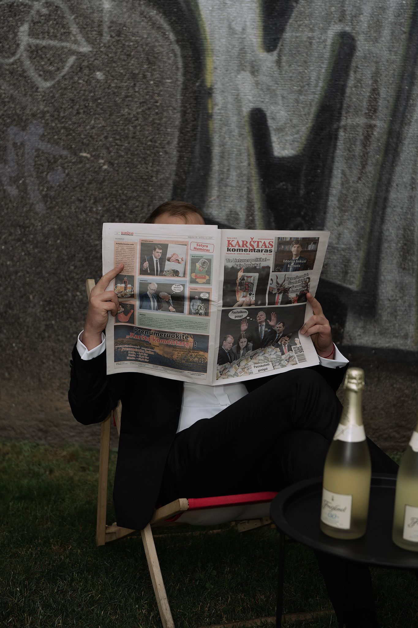 A person in formal attire reads a newspaper while sitting in a chair outdoors with a champagne bottle nearby.