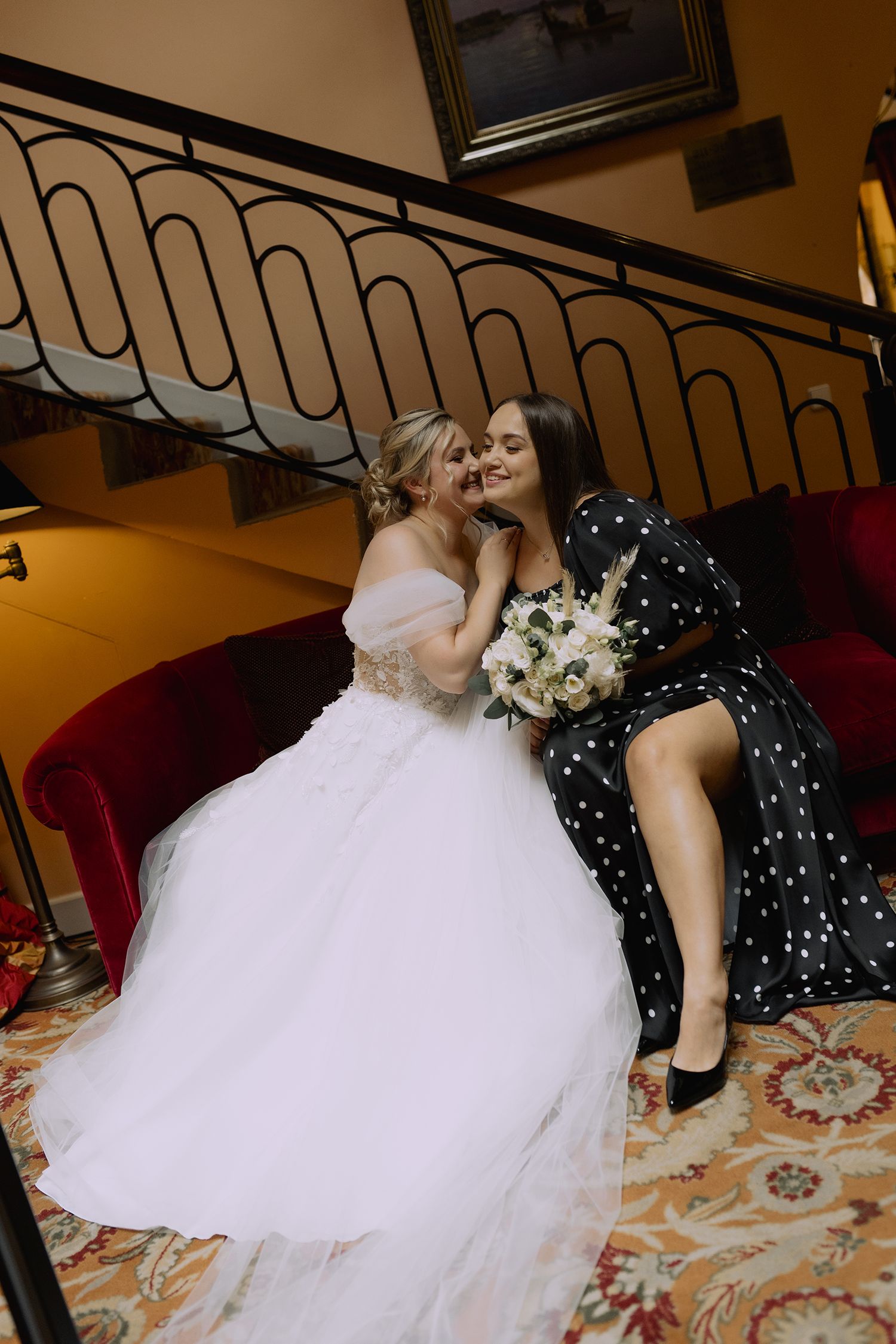 Two people share a tender moment on red velvet seating, one wearing a white ballgown and one in a polka dot dress.