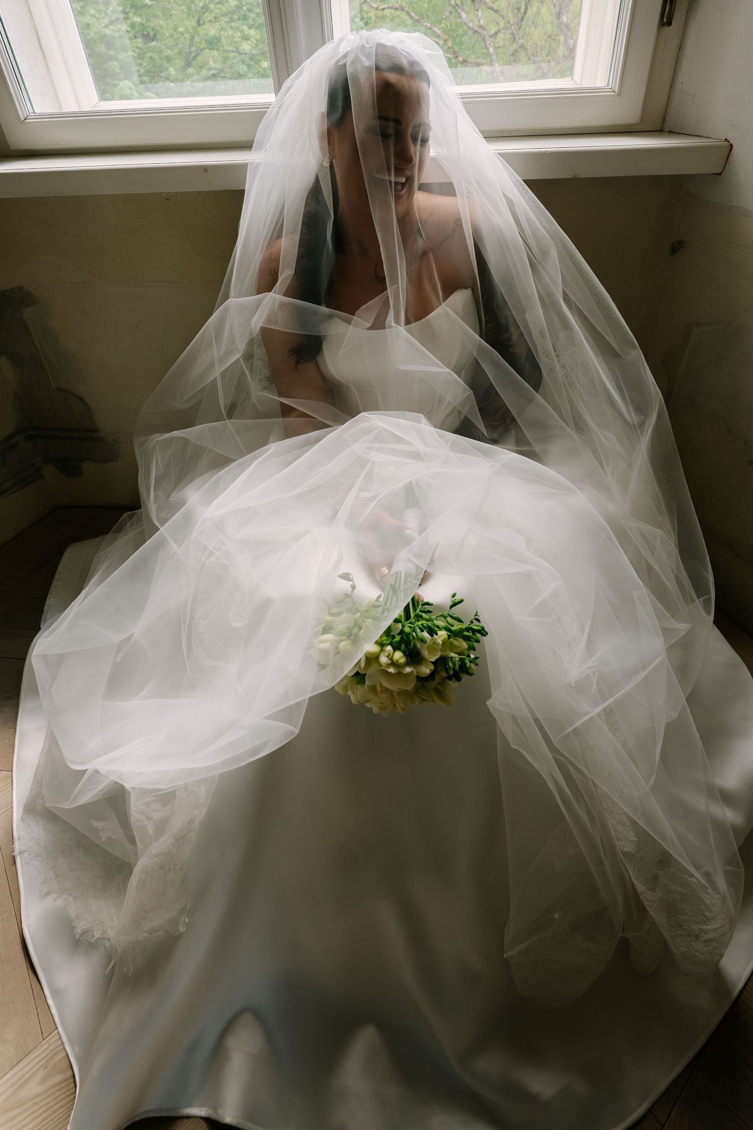 Bride in white gown and veil sits by window holding bouquet of white flowers in soft natural light.