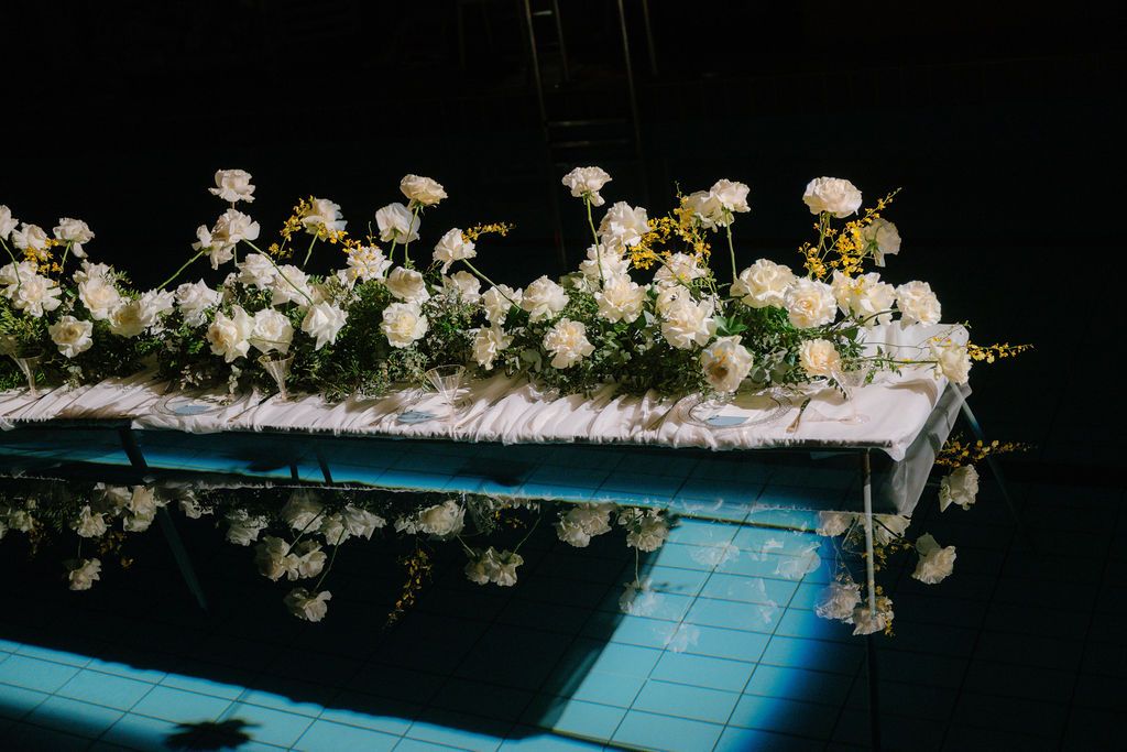 White roses and greenery arranged along a wedding aisle runner, reflected in the shimmering blue tiled water of a pool.