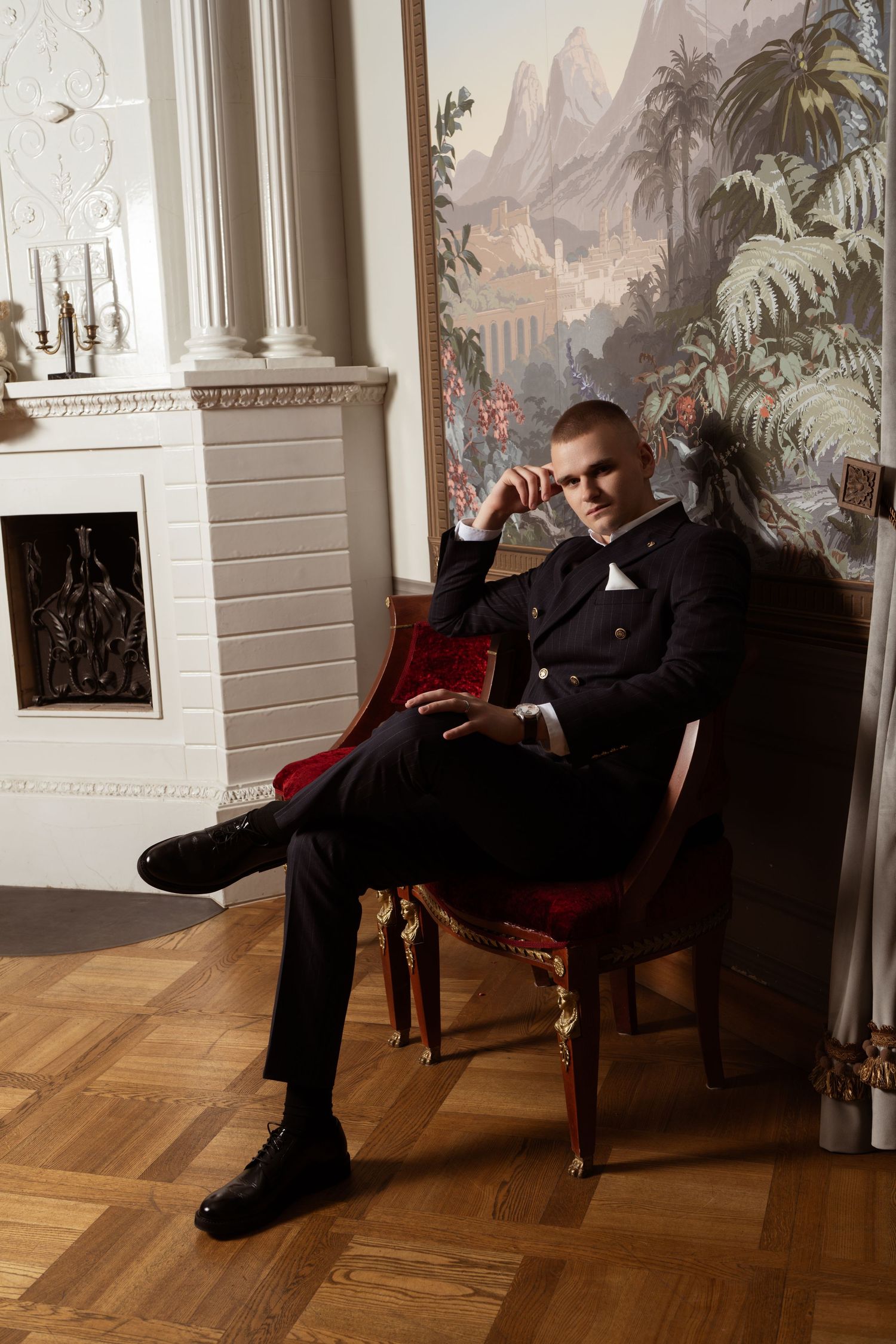 Man in a dark suit and white bow tie sits on a red velvet armchair in an elegant room with a fireplace and tropical mural.