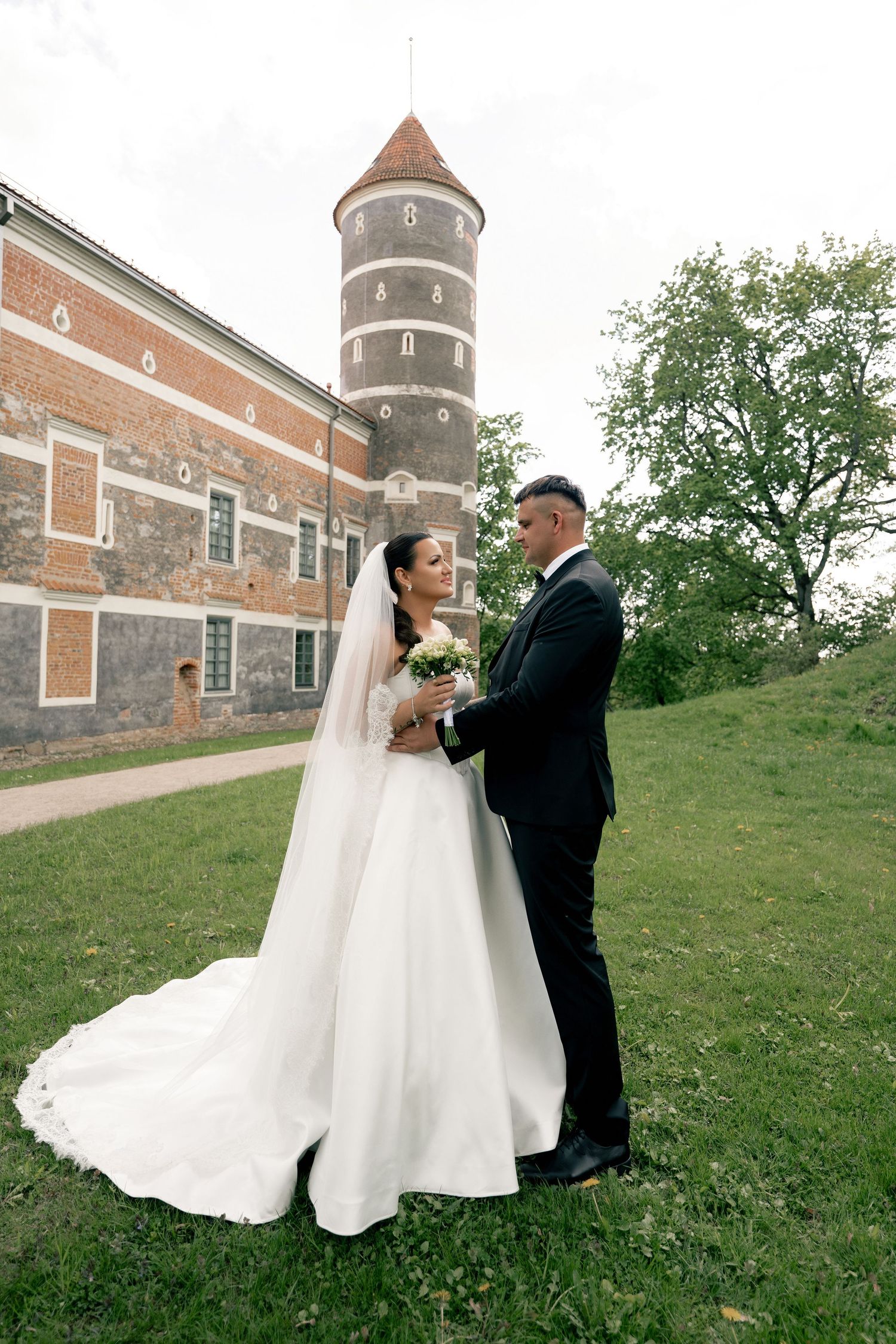 Bride and groom embrace on a grassy lawn beside a historic brick castle tower, the bride's long veil and train trailing behind.