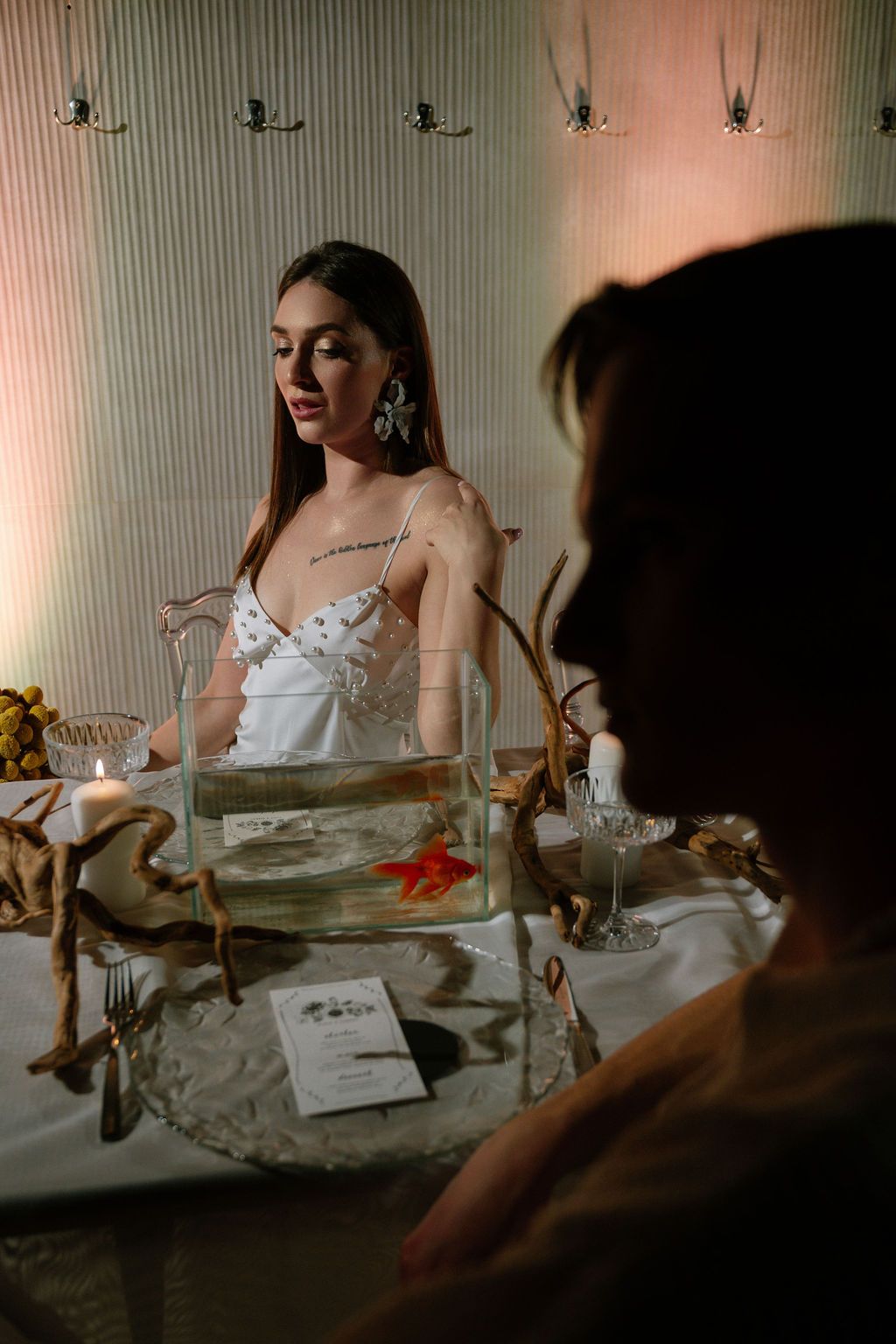 Woman in a white beaded dress sits at an elegant table with a goldfish bowl, driftwood branches, and a lit candle in warm light.