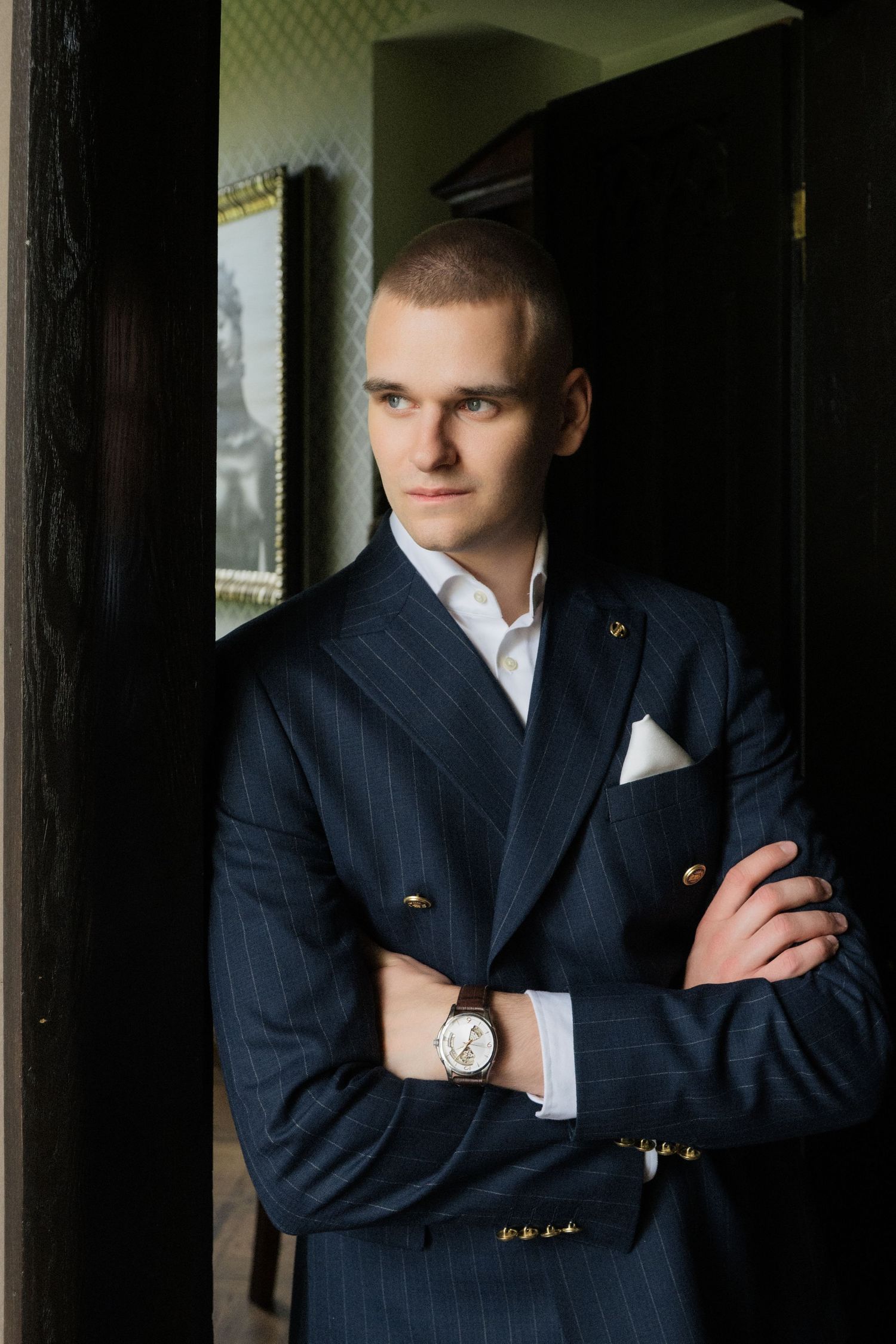 Man in a navy pinstripe double-breasted suit with gold buttons and a wristwatch stands with arms crossed in a dark doorway.