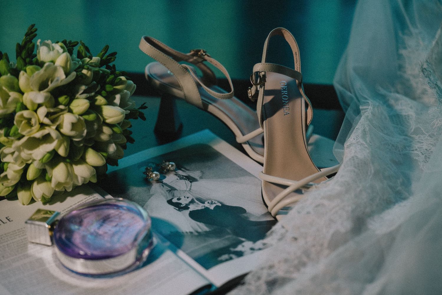 Elegant strappy heeled sandals beside a white floral bouquet, a compact mirror and wedding photos on a table with sheer lace fabric.