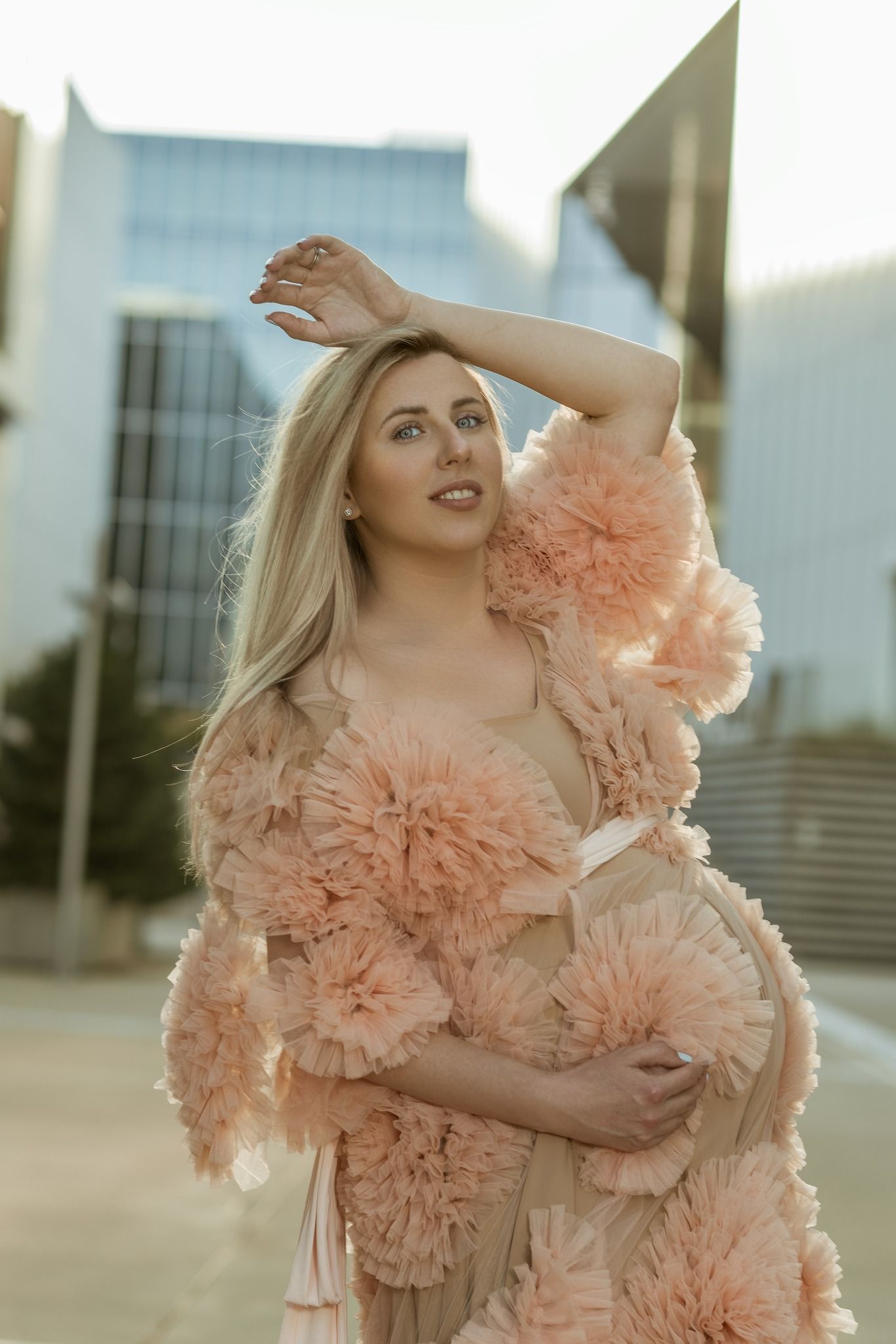 Pregnant woman in pink ruffled maternity gown posing outdoors with modern glass buildings in background.