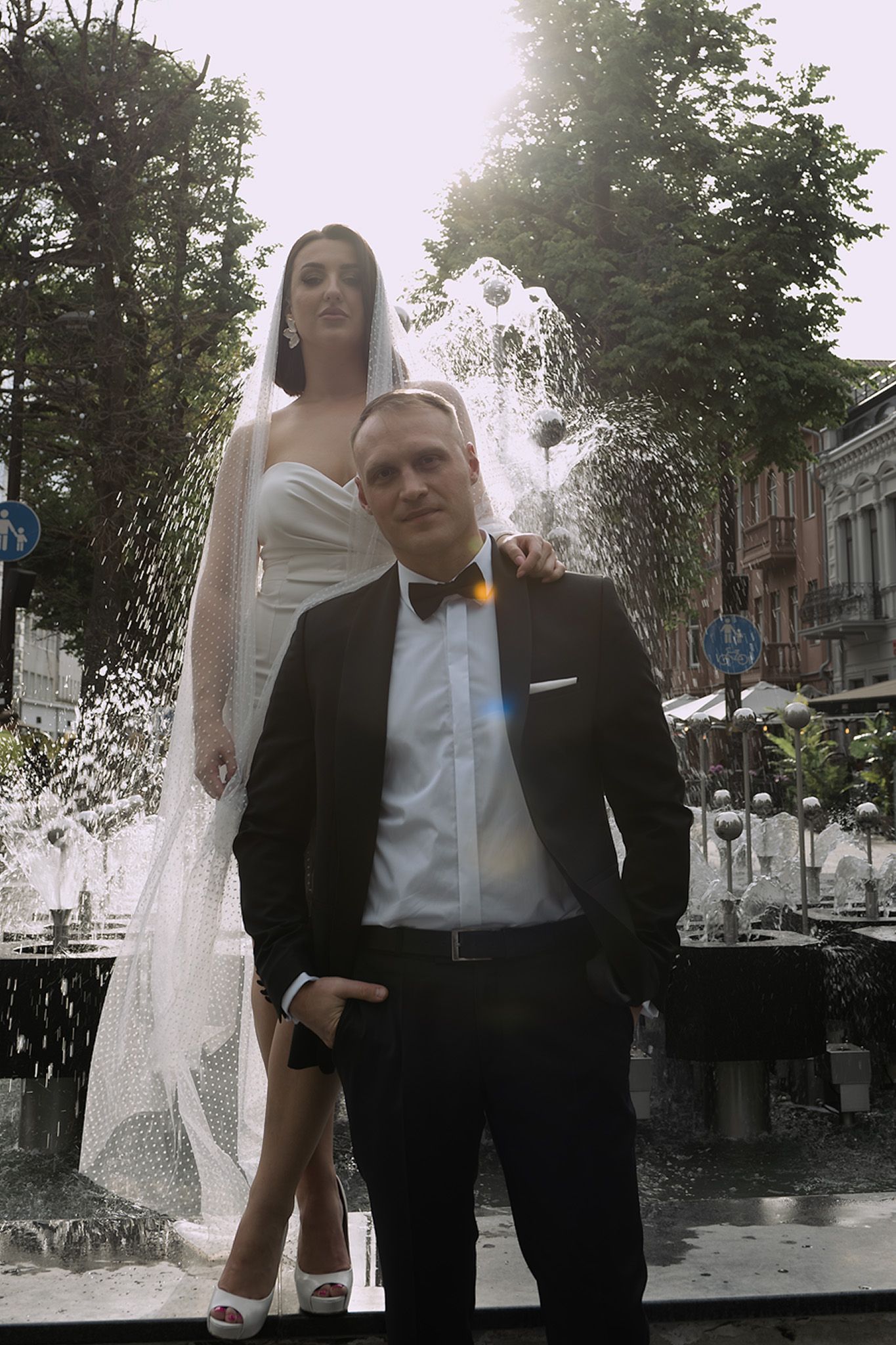 A bride and groom share a tender moment together outdoors under trees on a city street.