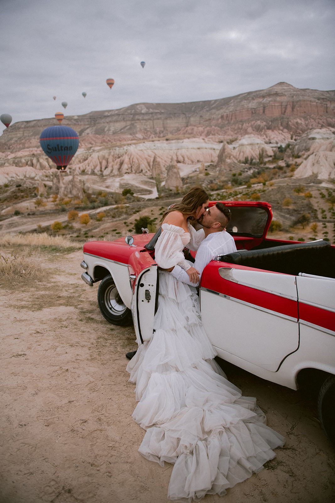 Hot air balloons float above desert cliffs as a classic convertible car and flowing white dress create a romantic scene in Cappadocia.