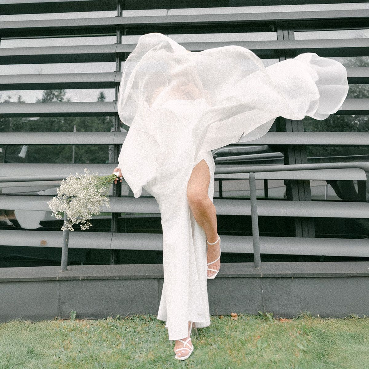 Close-up view of a bride's white dress and shoes on grass against a white bleacher backdrop.