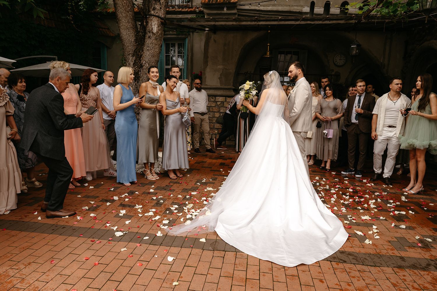 Wedding guests line a brick walkway tossing rose petals as a couple exits their ceremony in a grand departure.