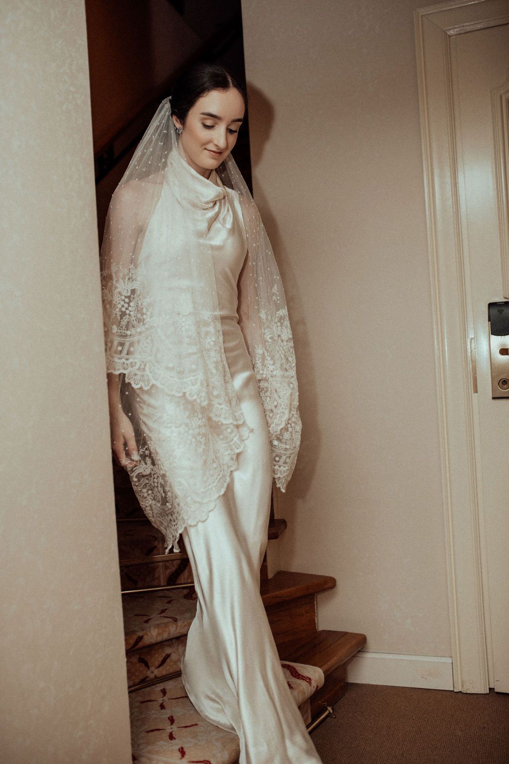 Bride in elegant ivory satin gown with lace veil descending staircase in soft warm lighting.