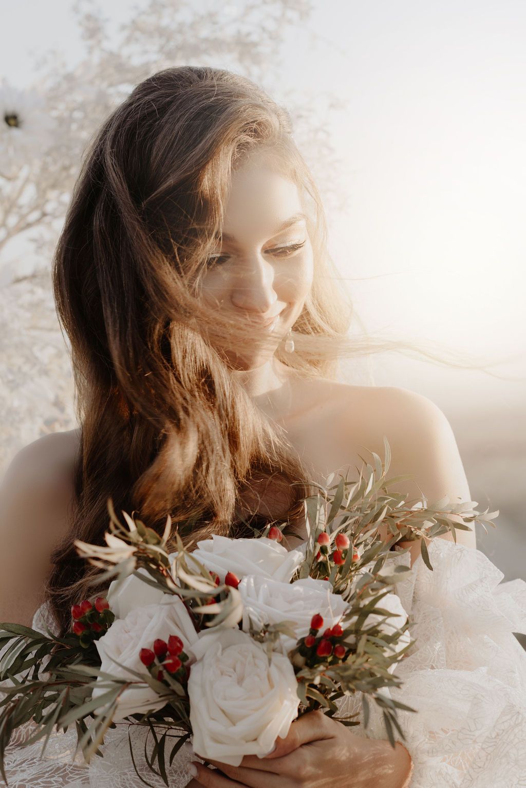 Radiant bride holding white roses and red berry bouquet smiles softly in golden sunlight.