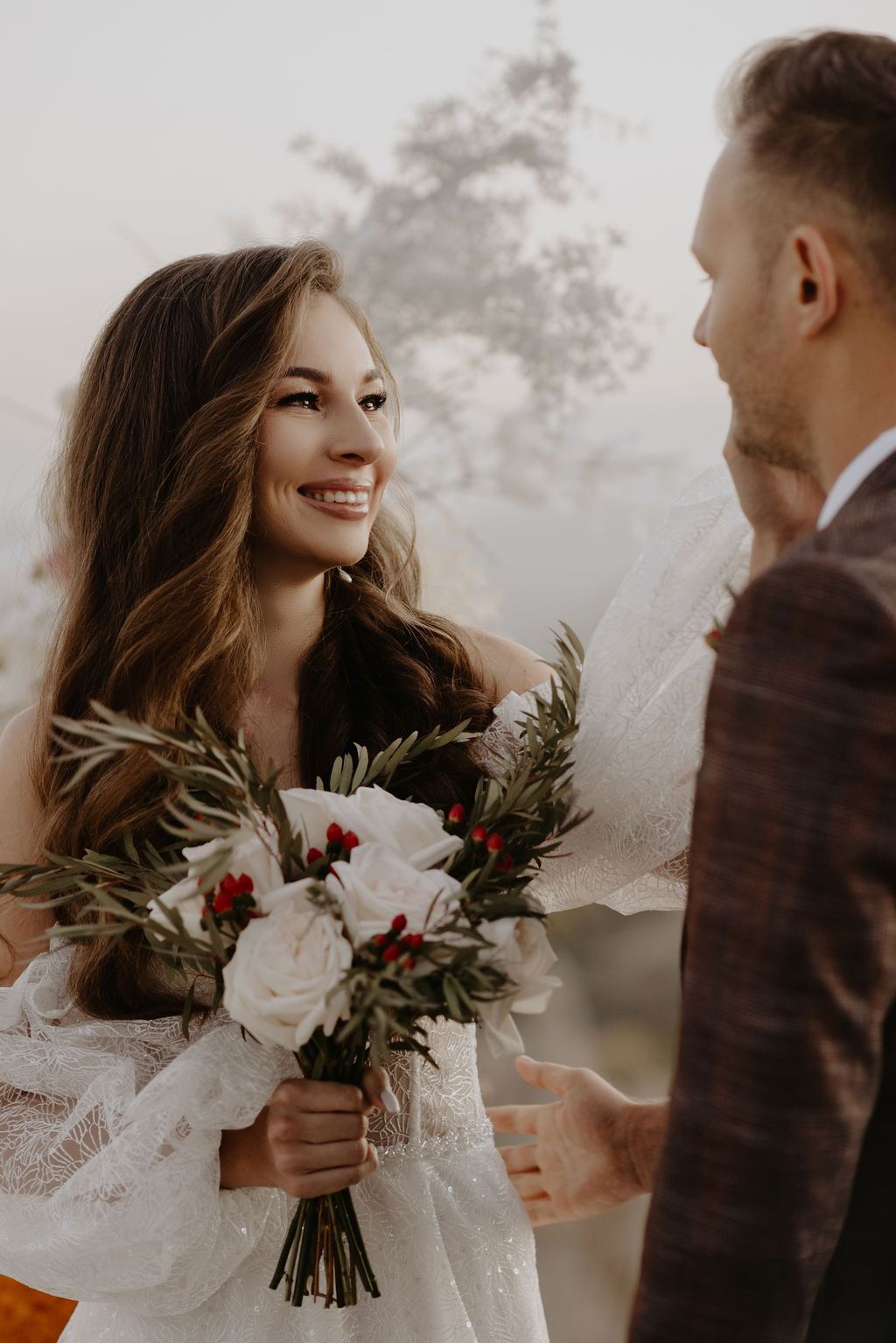 Smiling bride in lace gown holds white rose bouquet with red berries, gazing at groom in brown suit outdoors.