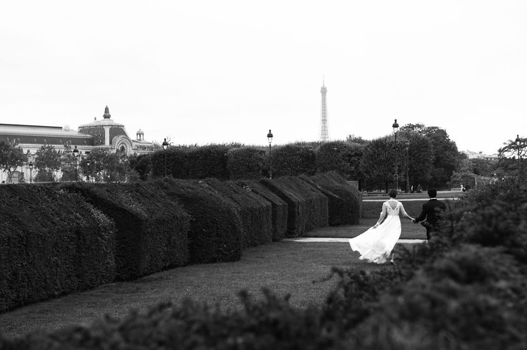 Bride in white gown walks through manicured hedges in Tuileries Garden with Eiffel Tower in black and white.