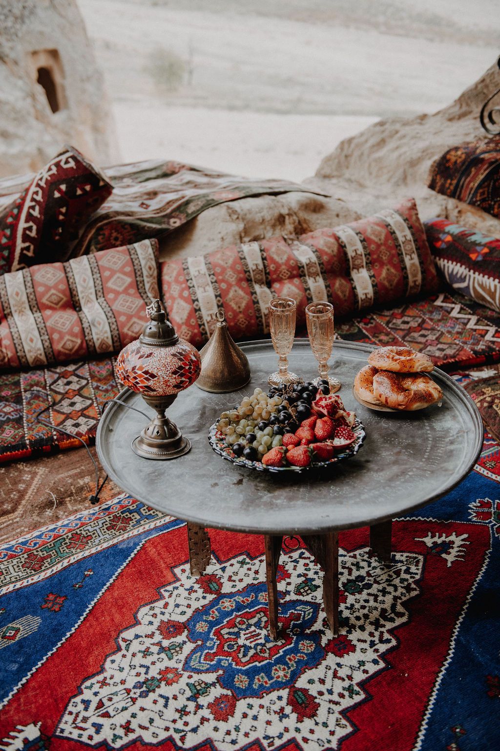 Bohemian outdoor setting with colorful Turkish kilim rugs, a low metal table with fresh fruits, champagne flutes, and bread.