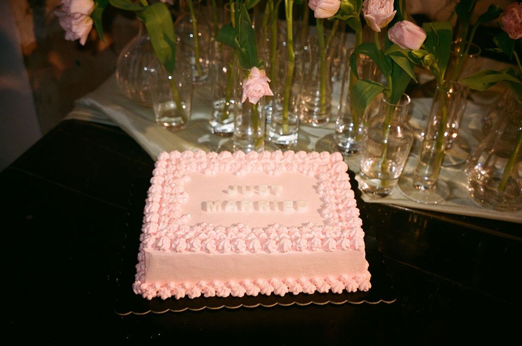 Pink square celebration cake with decorative frosting border displayed on table with roses and glassware.