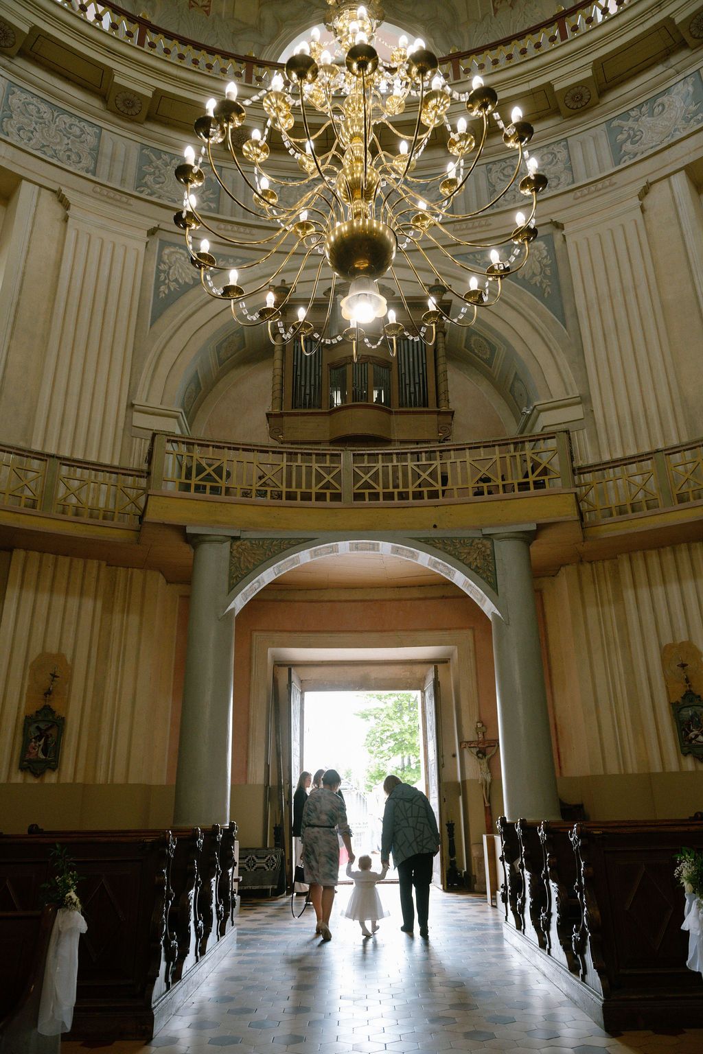 Elegant church interior with chandelier, organ loft, and family walking toward bright arched doorway.