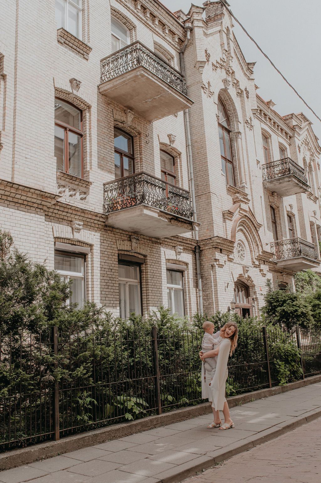 Woman holding a child on a sidewalk in front of an ornate white brick European building with balconies.
