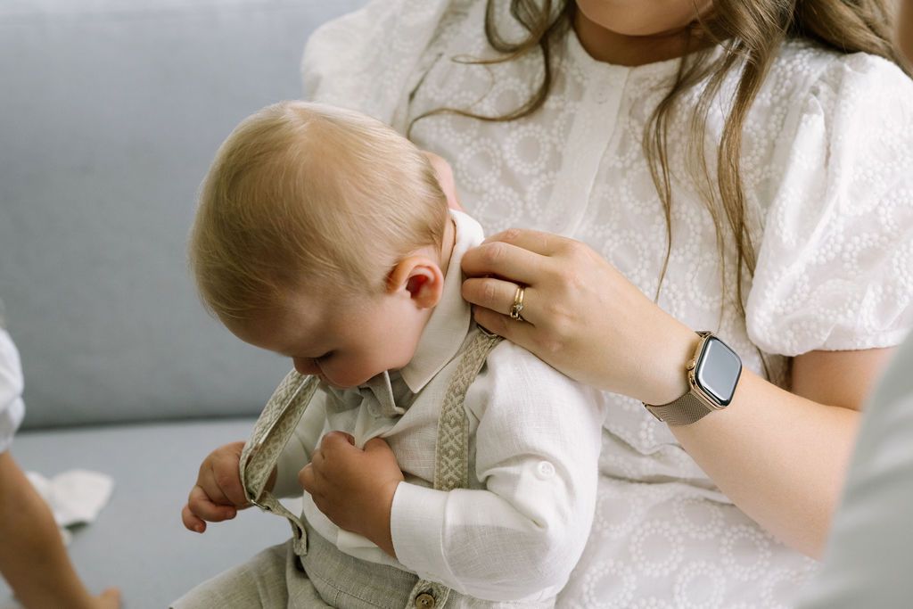 A mother in a white dress holds her baby boy in a white outfit on a gray sofa, adjusting his clothing.