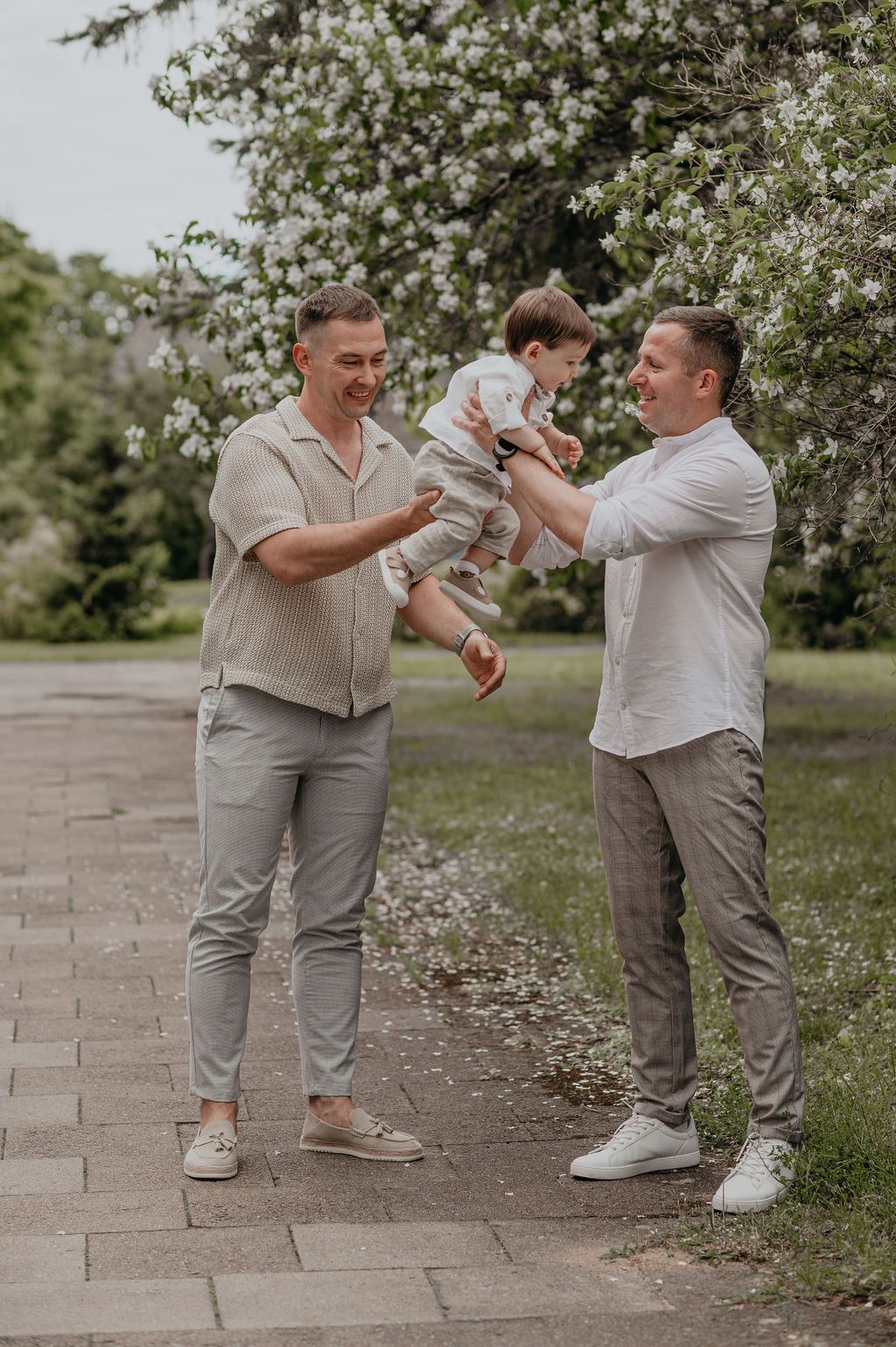Two men playing with a toddler outdoors near flowering trees on a paved path in a park.