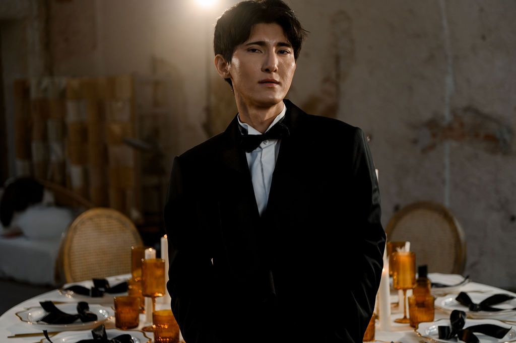 Elegant man in black tuxedo standing at a formal dinner table with orange centerpieces in a dimly lit venue.