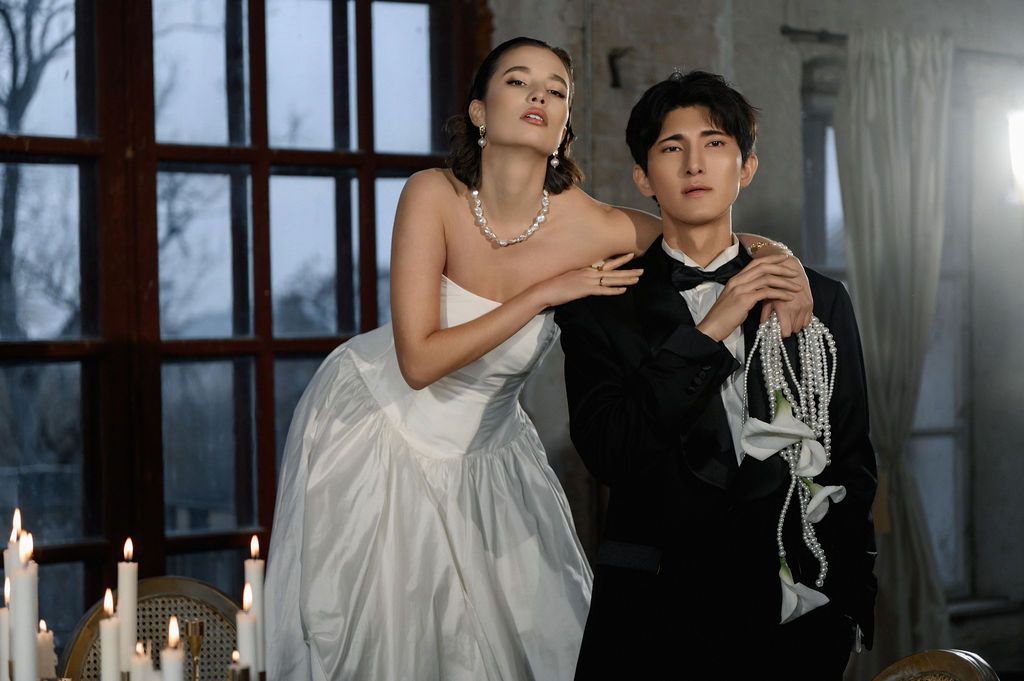 Elegant couple in wedding attire pose by candlelit window, woman in white gown with pearls, man in black tuxedo.