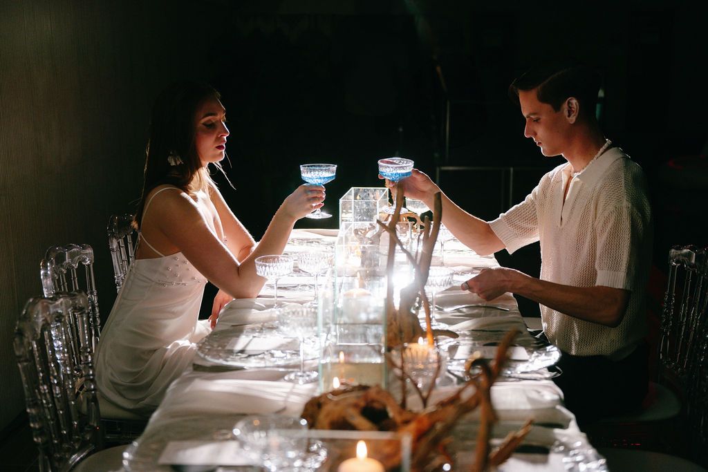 Couple toasting with glowing blue drinks at an elegant candlelit dinner table in a romantic dark setting.