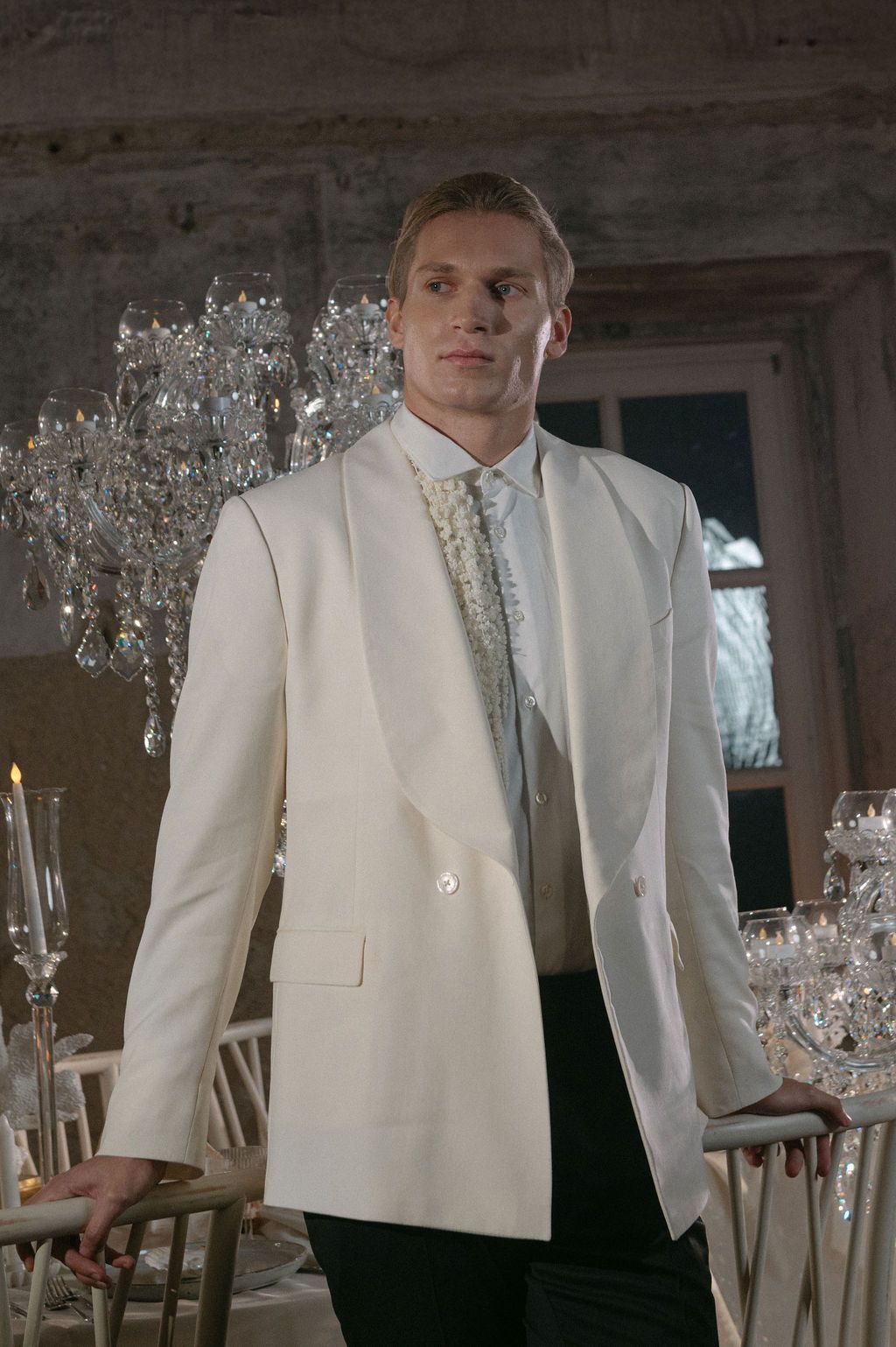 Man in white tuxedo jacket with decorative tie stands in elegant chandelier-lit venue.
