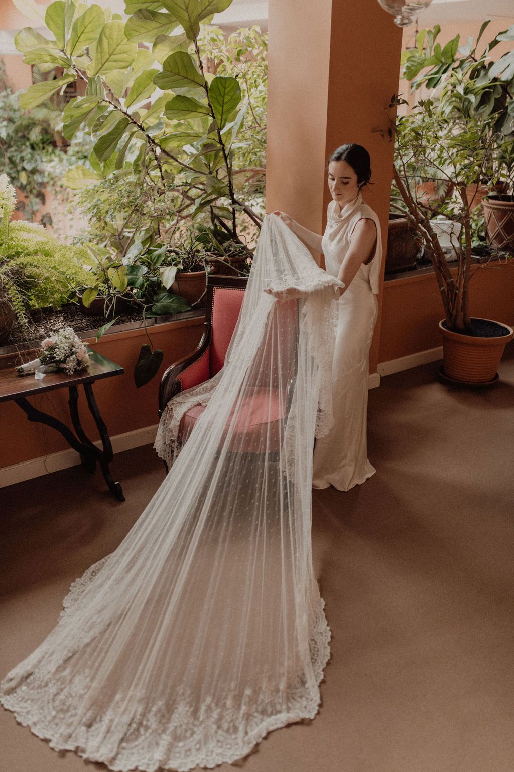 Bride in a white lace wedding gown holds a long cathedral veil surrounded by lush green plants indoors.