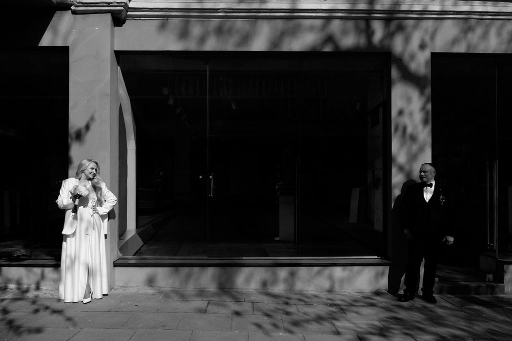 Black and white wedding photo of bride holding bouquet and groom in tuxedo standing apart outside a building.