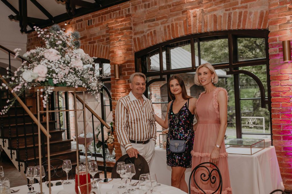 Three elegantly dressed guests posing together at a decorated wedding reception venue with brick walls and floral arrangements.