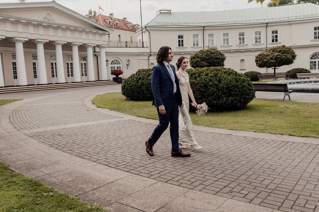 A couple walks hand in hand on a brick path outside an elegant white neoclassical building with manicured hedges.