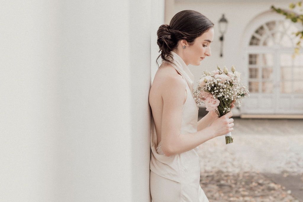 Elegant bride in sleeveless gown holds a delicate pink and white bouquet while leaning against a white pillar.