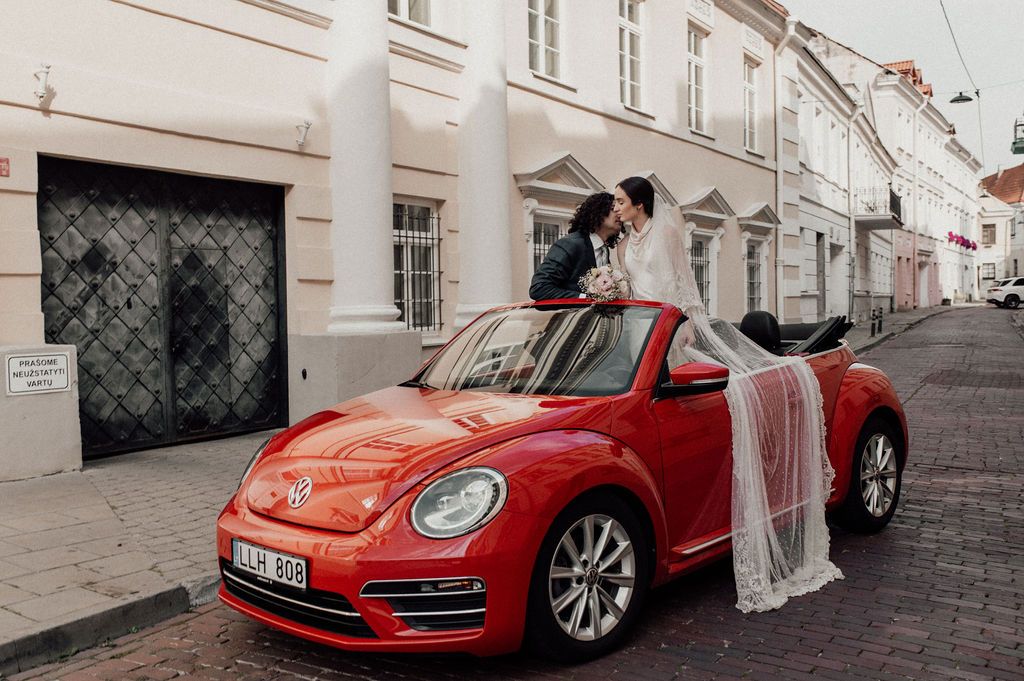 Bride and groom kissing in a red VW Beetle convertible on a cobblestone street.