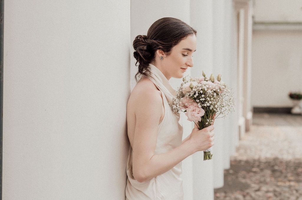 Bride in elegant dress smelling a delicate white and pink bouquet while leaning against a white column.