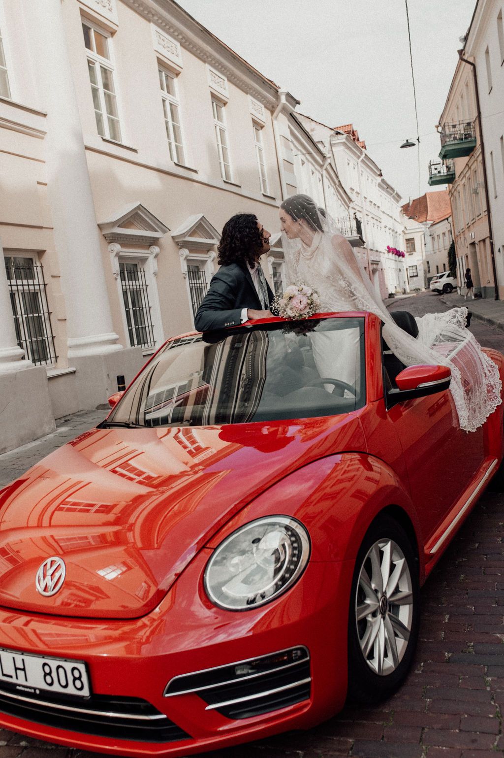 Bride and groom sharing a romantic kiss atop a red Volkswagen Beetle convertible on a European cobblestone street.