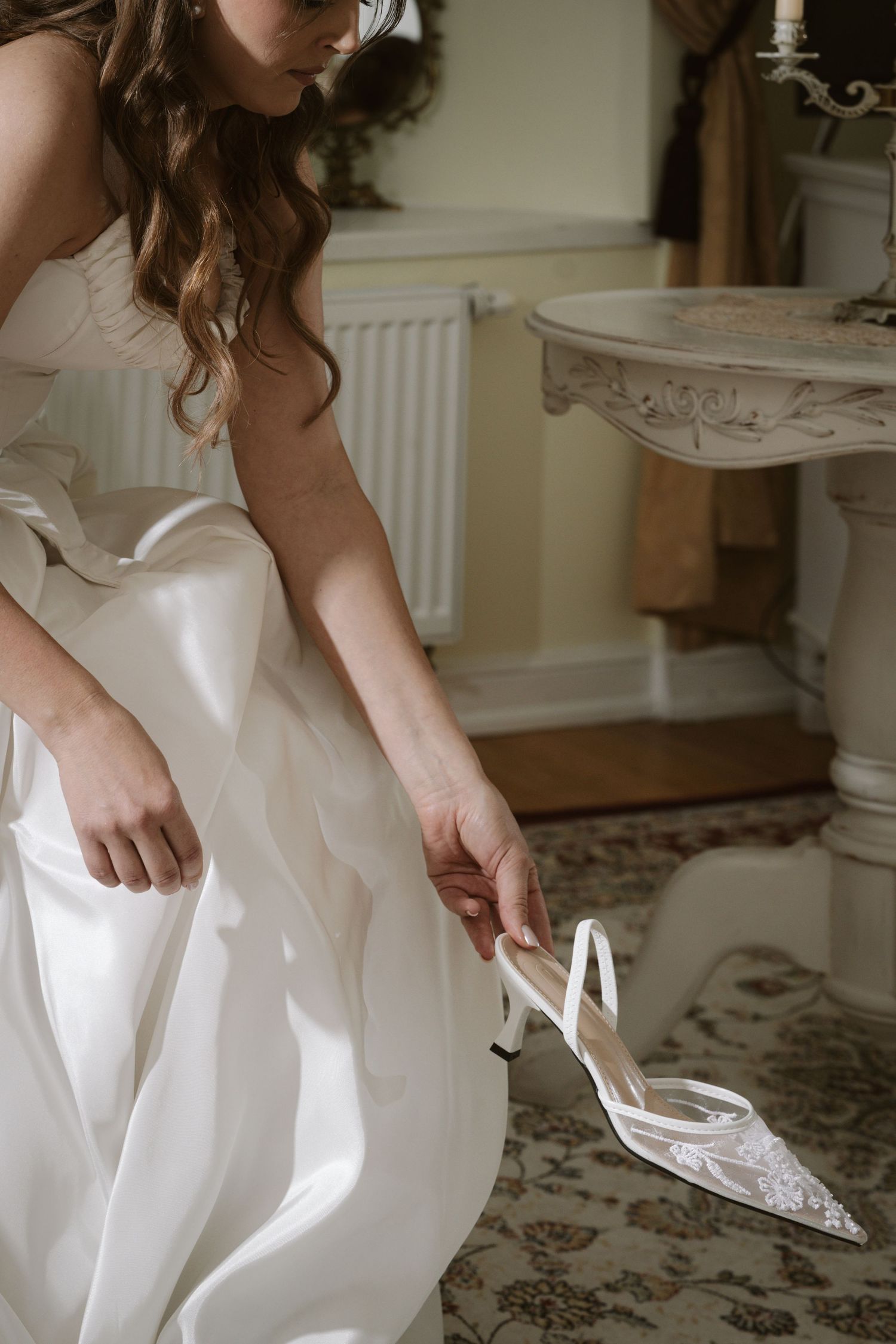 Bride in white satin gown holding elegant pointed-toe kitten heel wedding shoe in ornate vintage room.