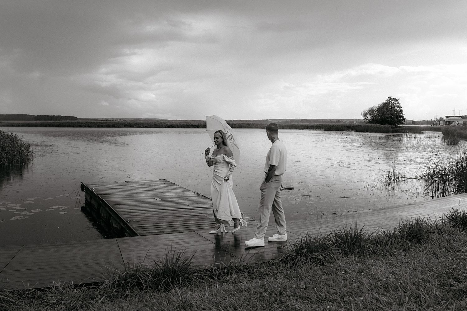 Two people in white clothing stand on a grassy bank beside a wooden dock on a serene lake in black and white.
