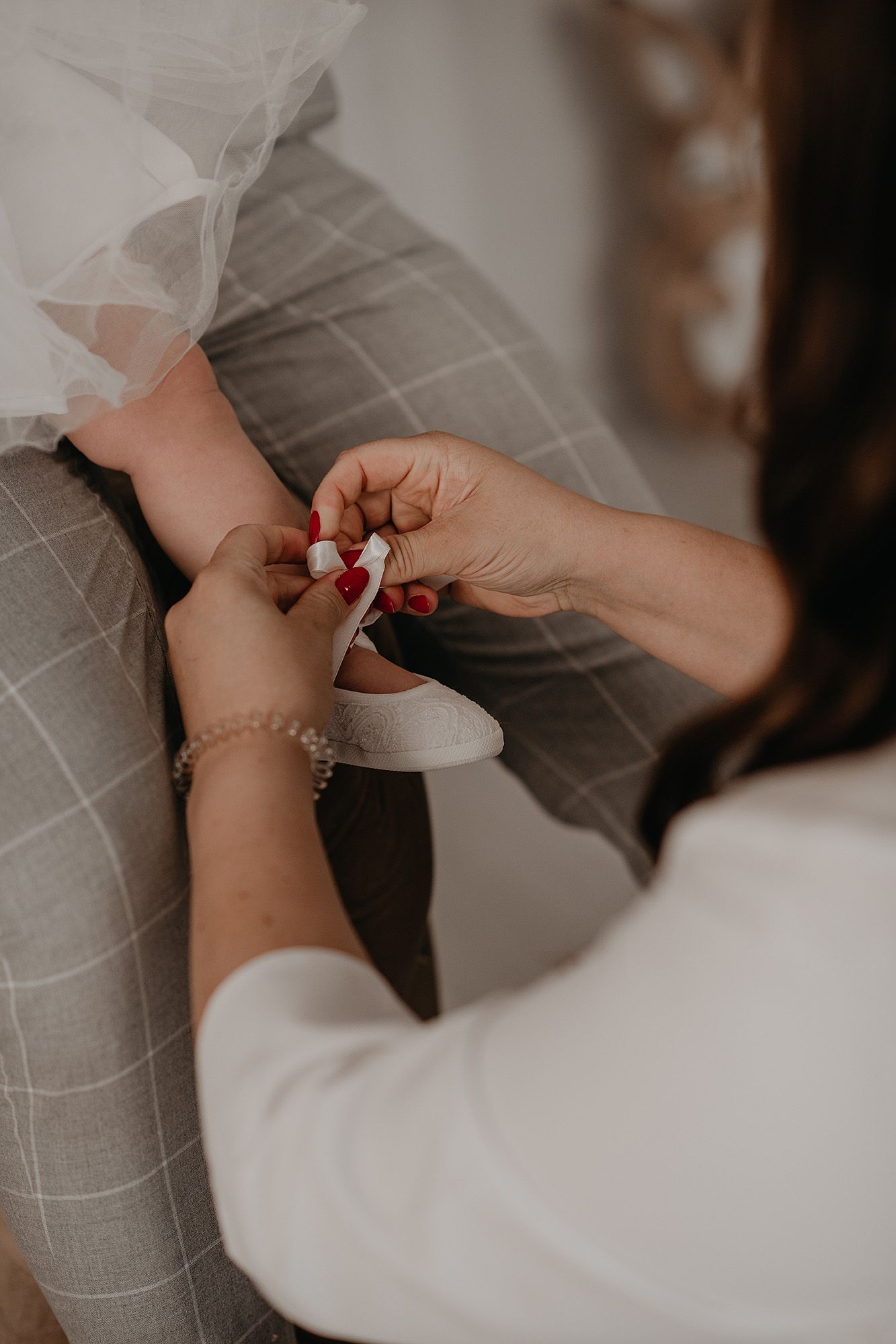 Close-up of hands with red nail polish fastening a delicate white wedding dress button against a gray background.