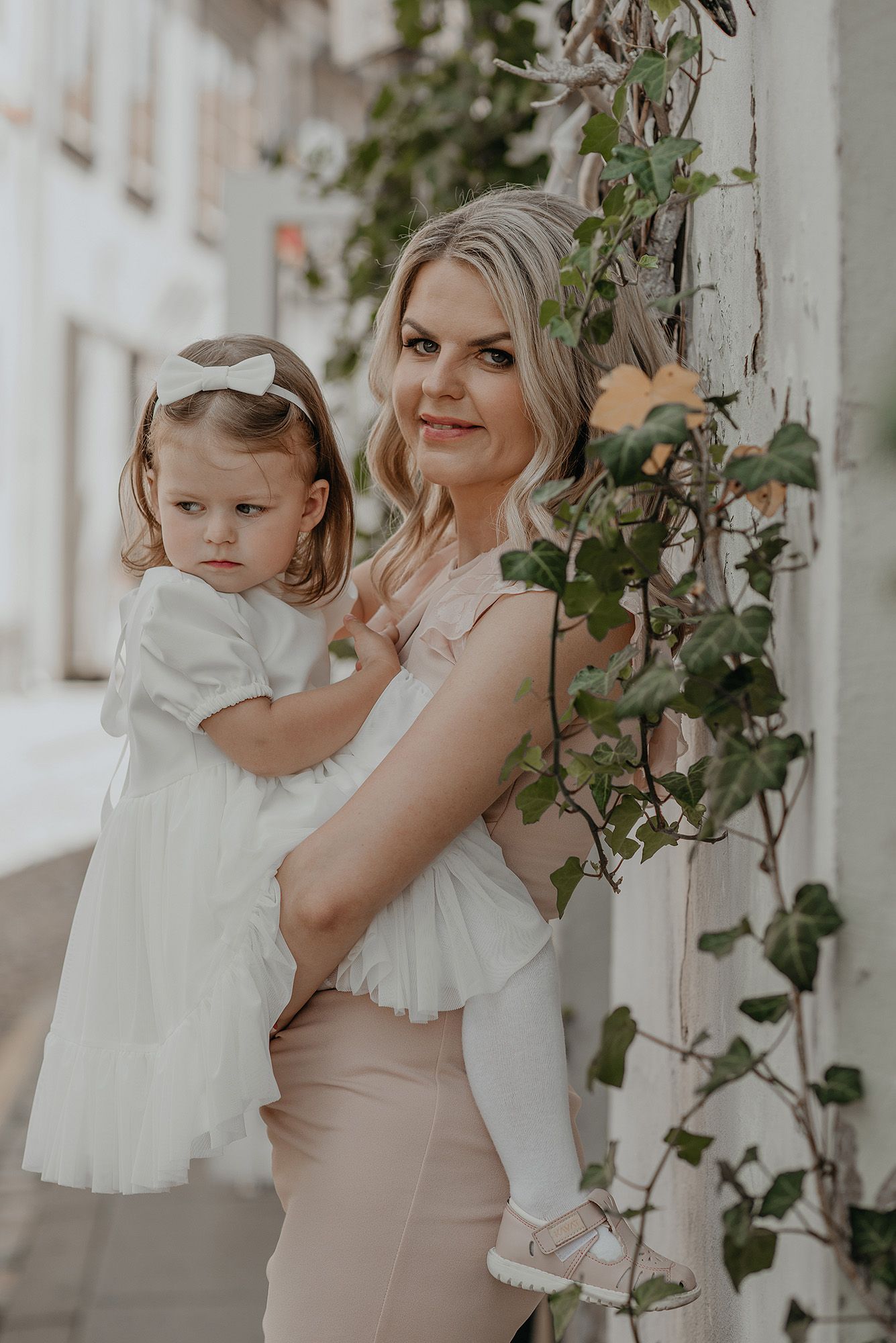 Mother and daughter in white dresses pose together with climbing vines on a white porch in a tender moment.