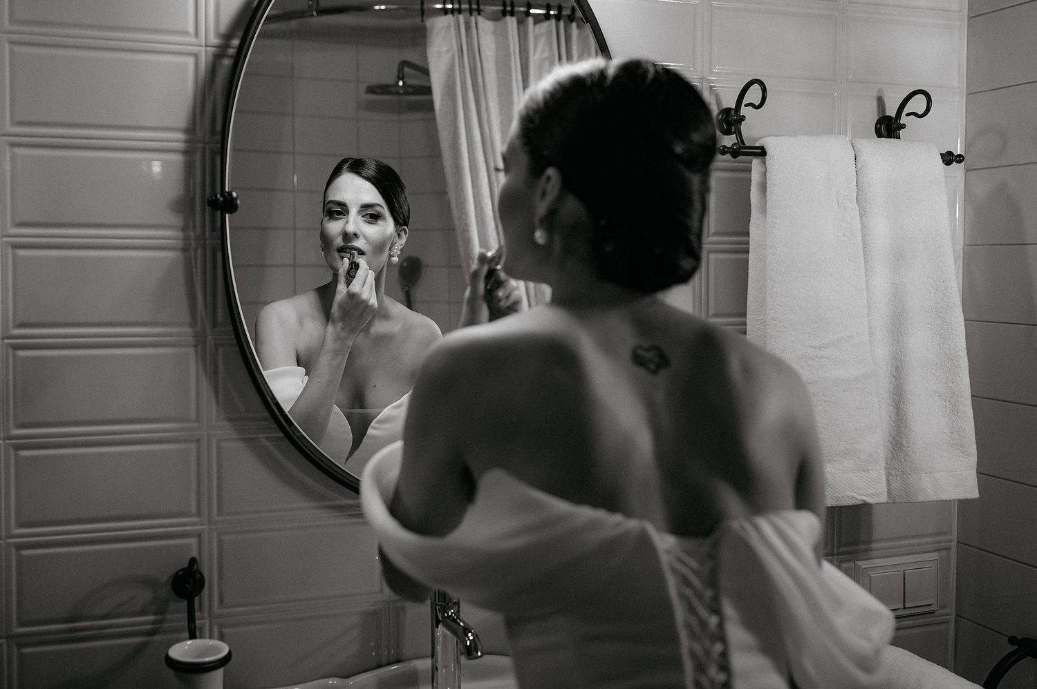 Black and white photograph showing a reflection in a bathroom mirror with someone applying makeup in an intimate moment.