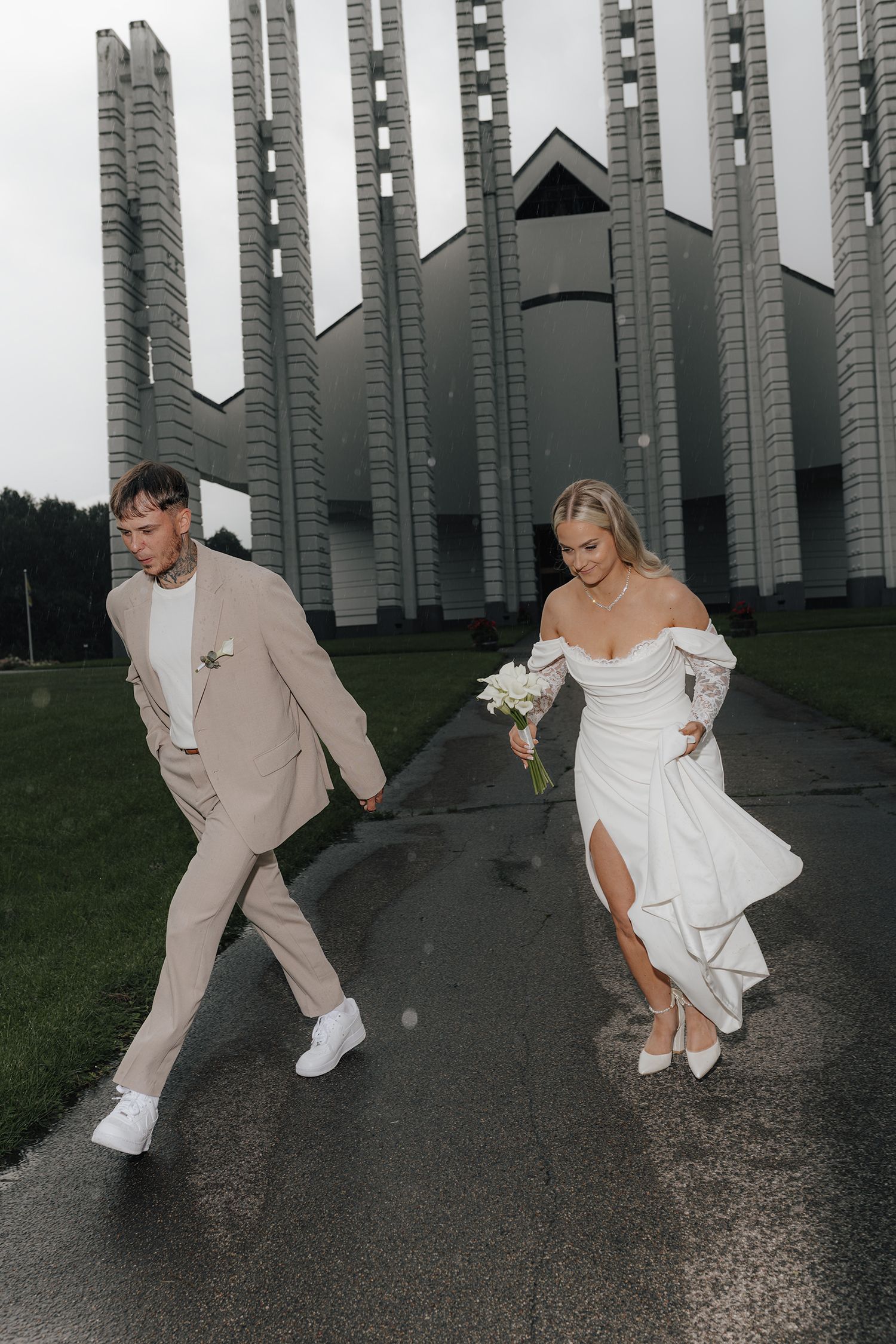 A couple in wedding attire playfully poses in front of tall concrete pillars at dusk.