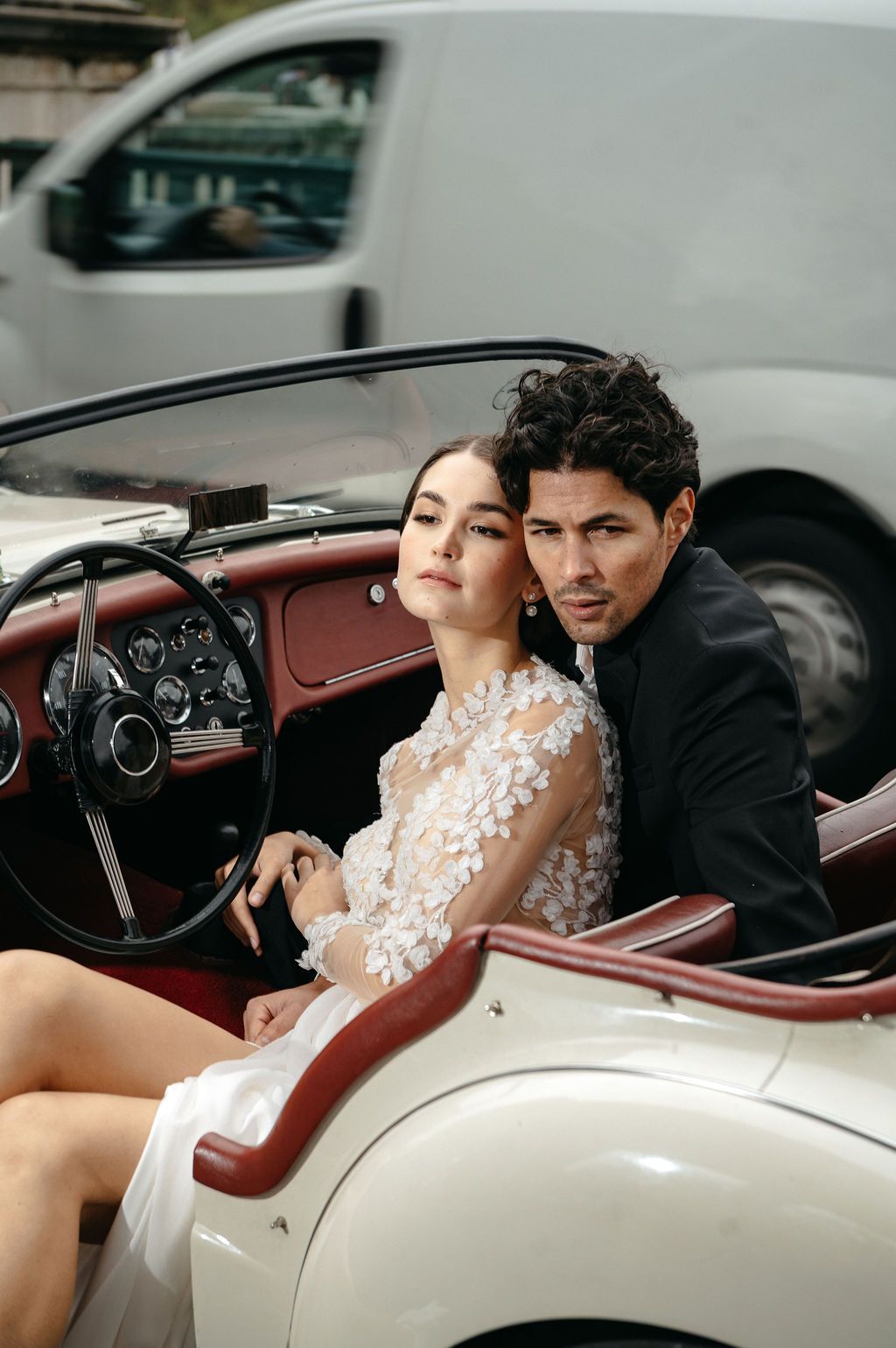 Elegant couple in wedding attire posing in a vintage white convertible car, bride wearing floral lace gown.